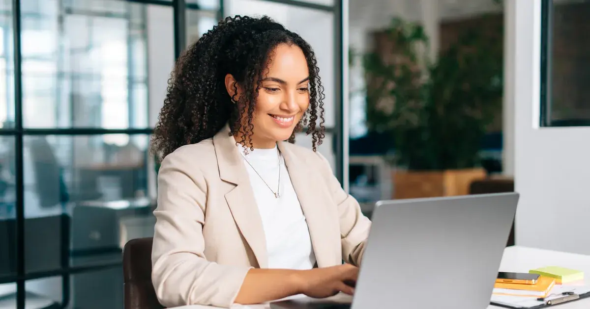 Finance professional in beige blazer smiling while working on expense reports and financial documents on a laptop at a desk in a modern office